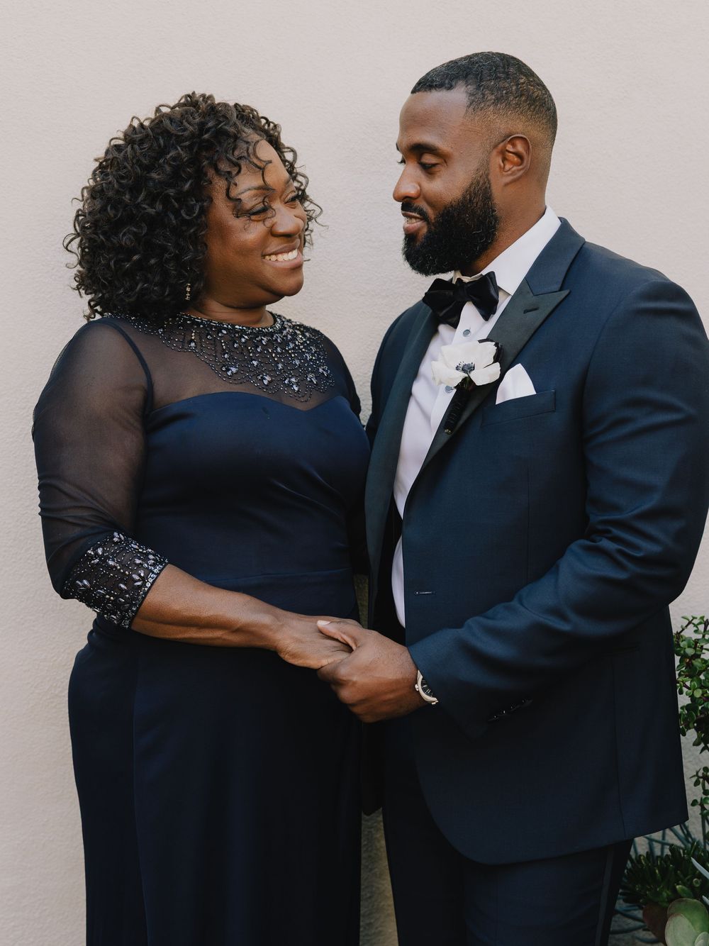 A groom in a navy tuxedo and his mother in a navy gown share a joyful moment at a wedding.