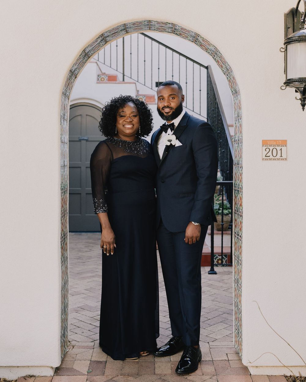Groom and older woman pose together in navy formal wear under stone archway.