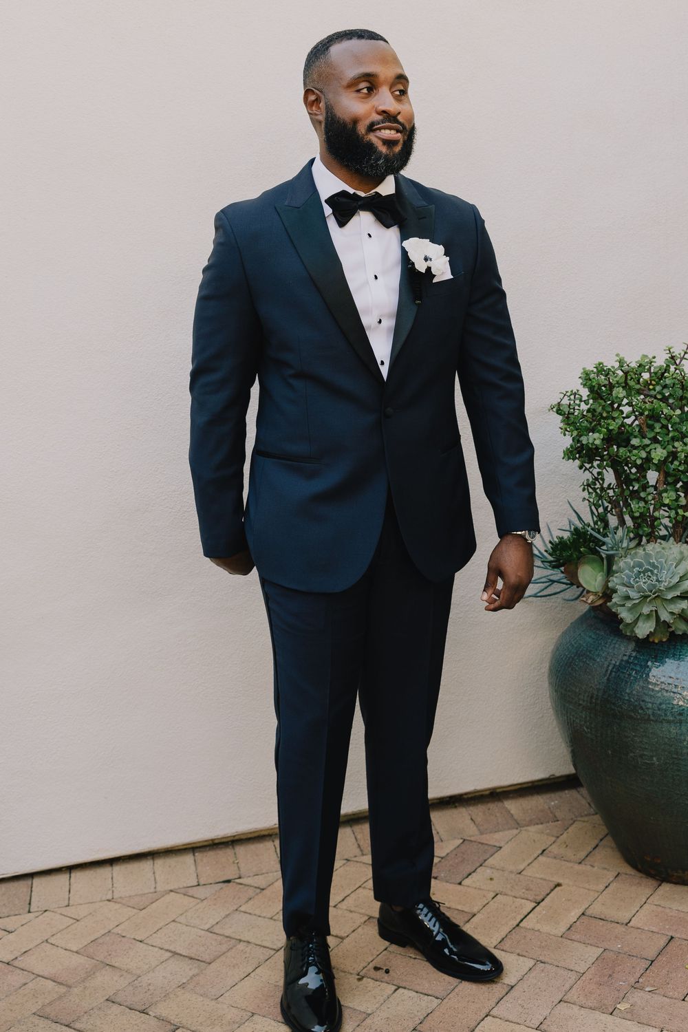 Dapper groom stands confidently in a navy tuxedo with black bow tie and white boutonniere outdoors.