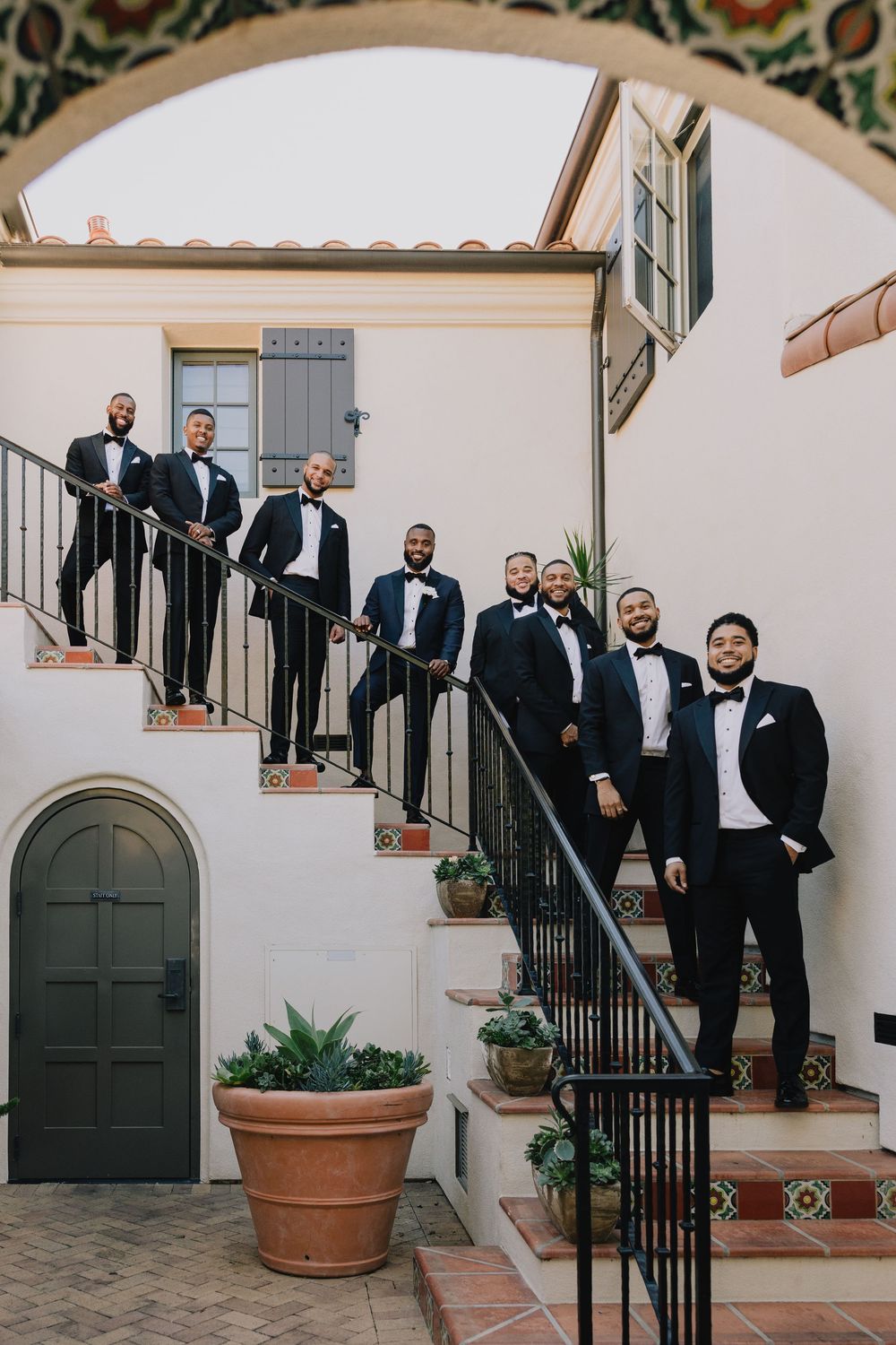 Groom and groomsmen in black tuxedos posed on outdoor staircase at Spanish-style wedding venue.
