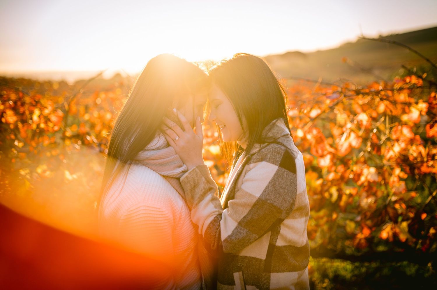 Two women sharing an intimate moment in a sunlit autumn vineyard with warm golden light and colorful fall foliage.