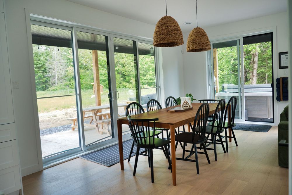 Modern dining room with wooden table, black chairs, pendant lights, and large glass doors.