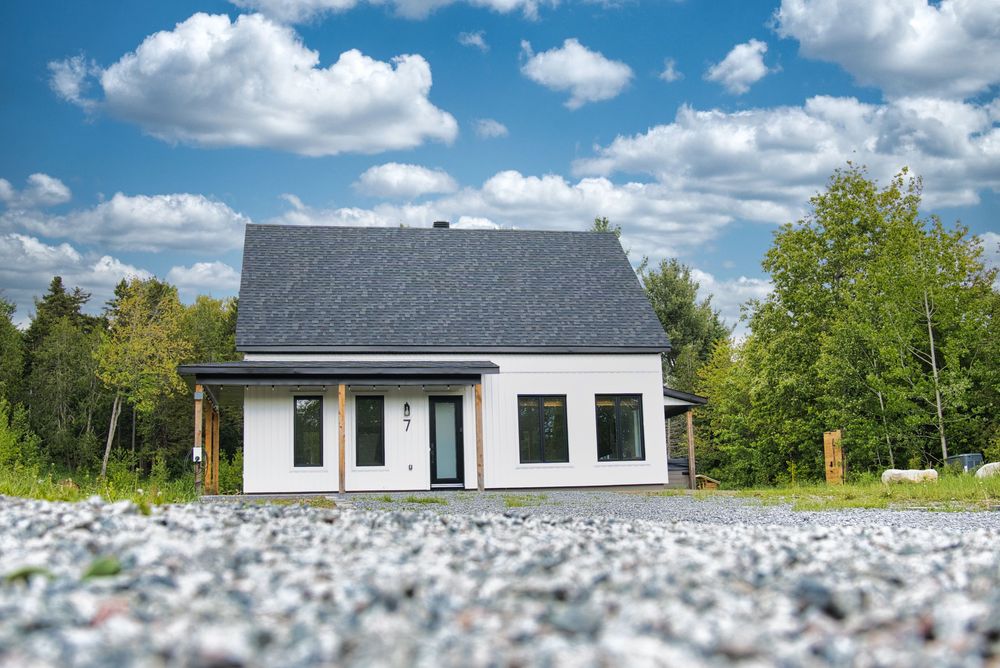 Modern white house with dark roof surrounded by lush trees under blue sky with white clouds.