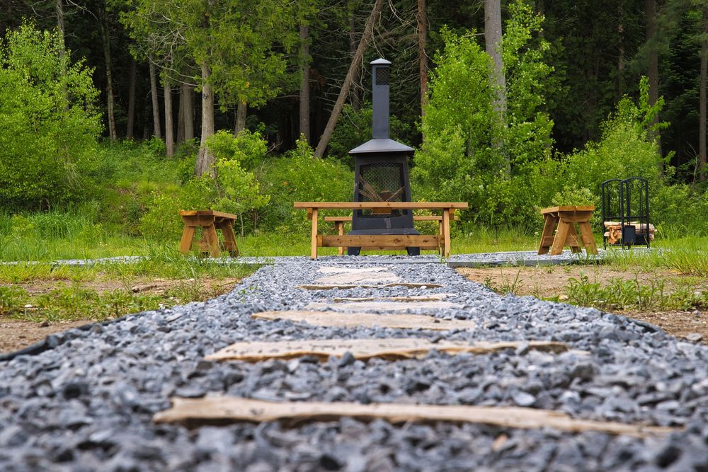 Outdoor fireplace with wooden benches on gravel path surrounded by green forest.