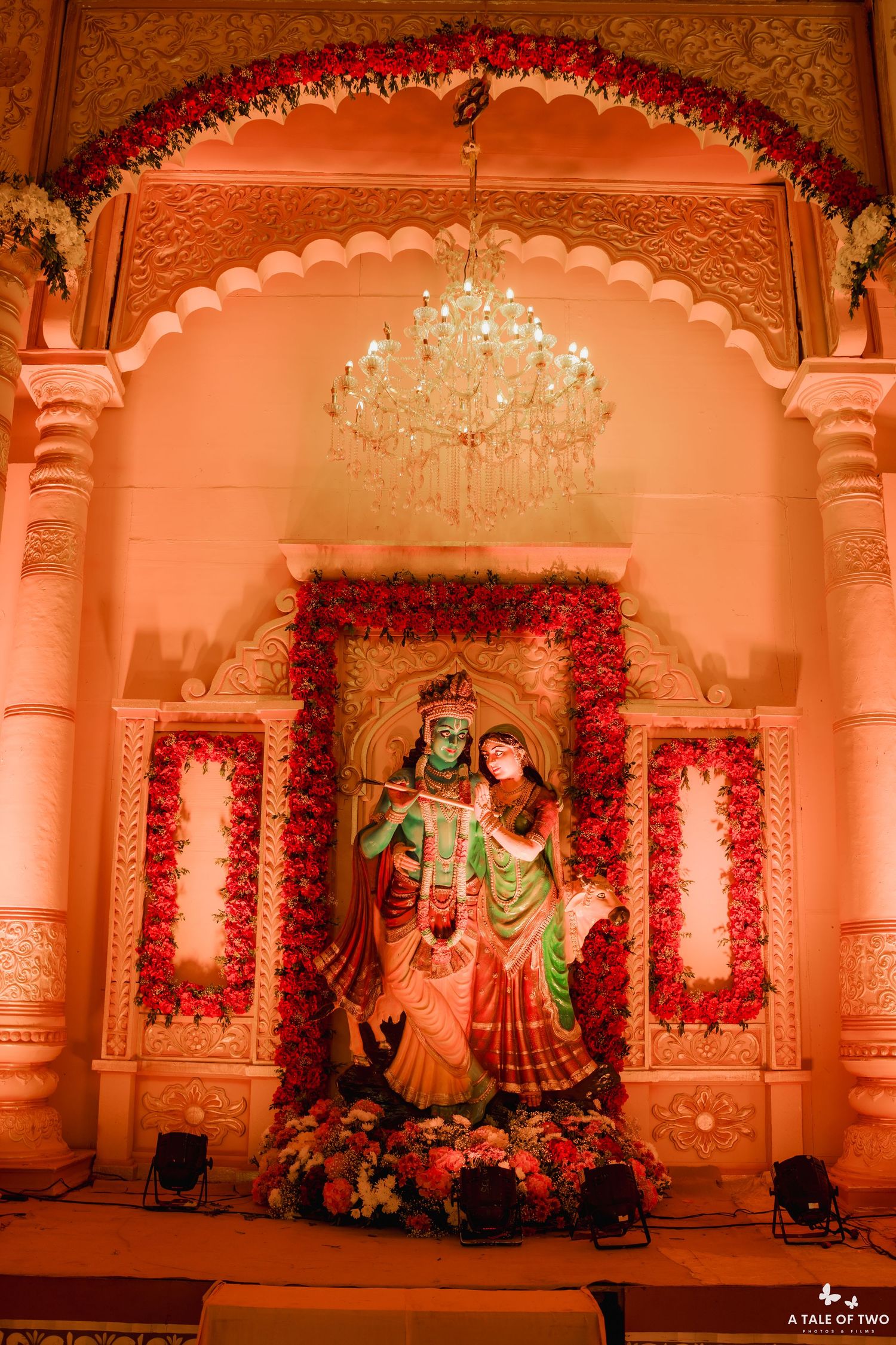 Radha Krishna idol adorned with floral garlands displayed under a crystal chandelier at an Indian wedding mandap.