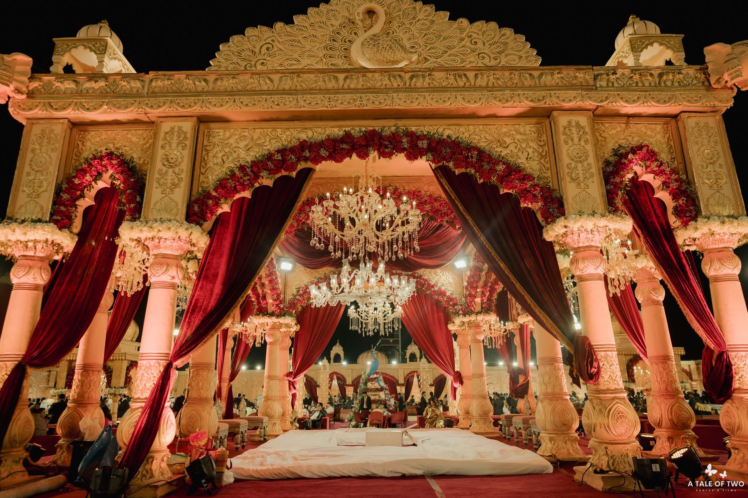 Ornate Indian wedding mandap with red drapes, crystal chandeliers, floral decor, and grand white archway illuminated at night.