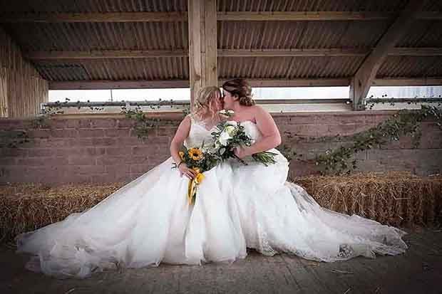 Two brides in white ball gowns holding bouquets pose romantically in a rustic barn with hay bales.