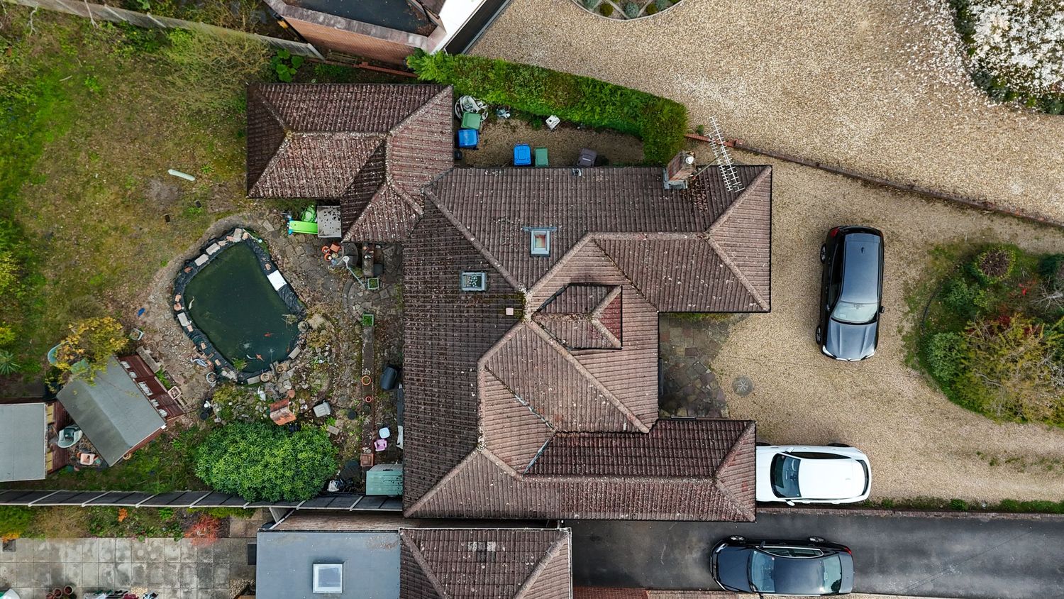 Aerial drone view of a residential house with brown tile roof, parked cars, and a small garden pond.