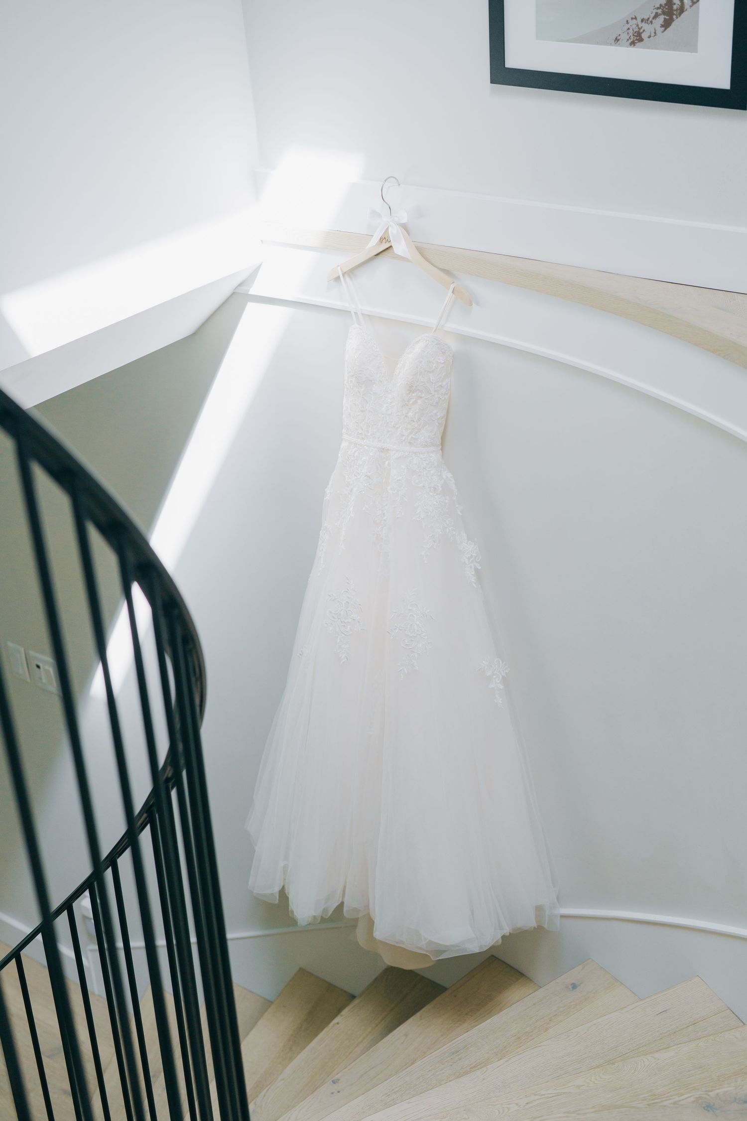 A white wedding dress hangs elegantly against a white wall near a black spiral staircase in natural light.Queen Elizabeth Park Wedding