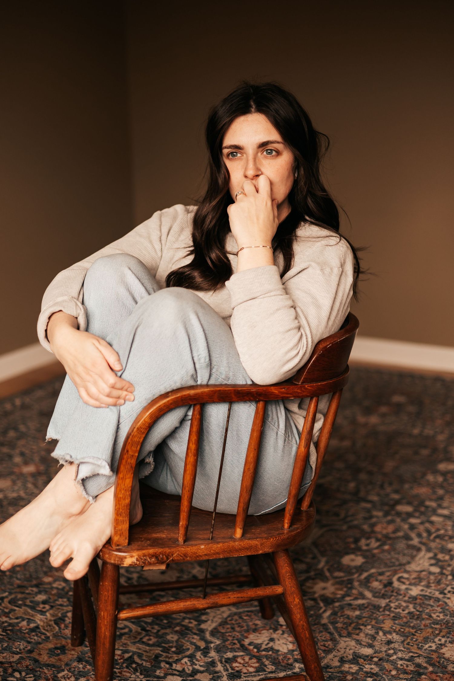 Woman in casual gray outfit sitting barefoot on wooden chair, hand near face, thoughtful expression  |My Postpartum Depression Story
