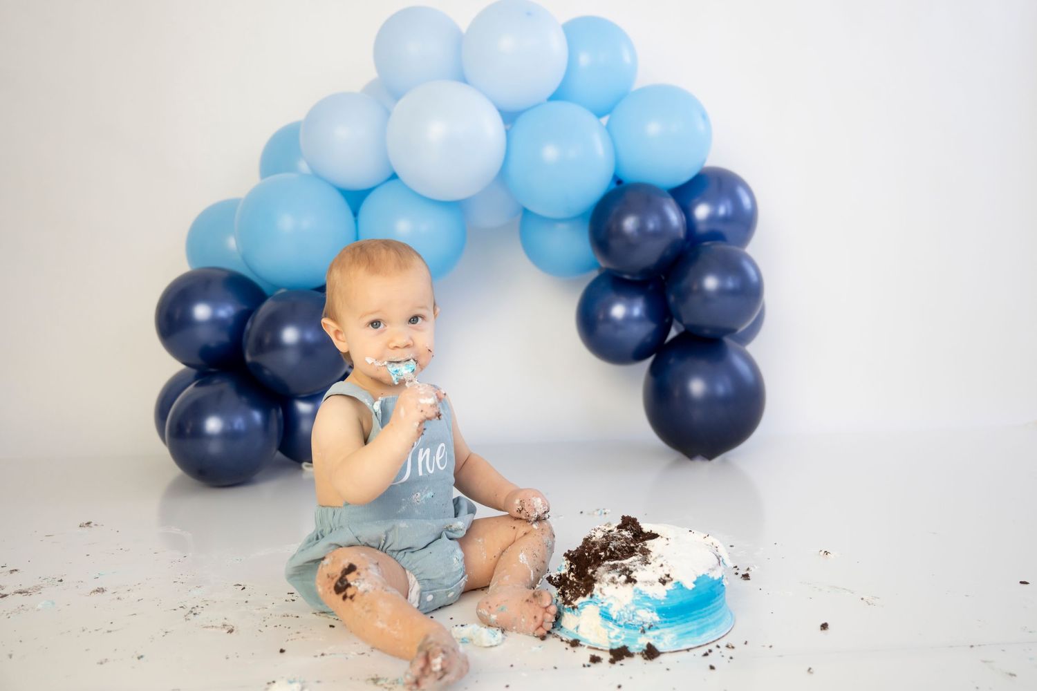 Baby in diaper enjoying cake smash photo session with blue and navy balloon arch decorations and confetti.