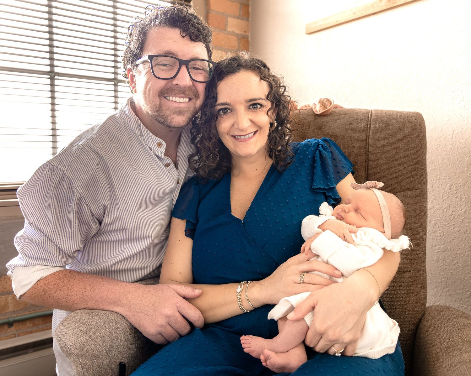 A couple in casual attire sits on a couch holding a newborn baby while smiling at the camera.