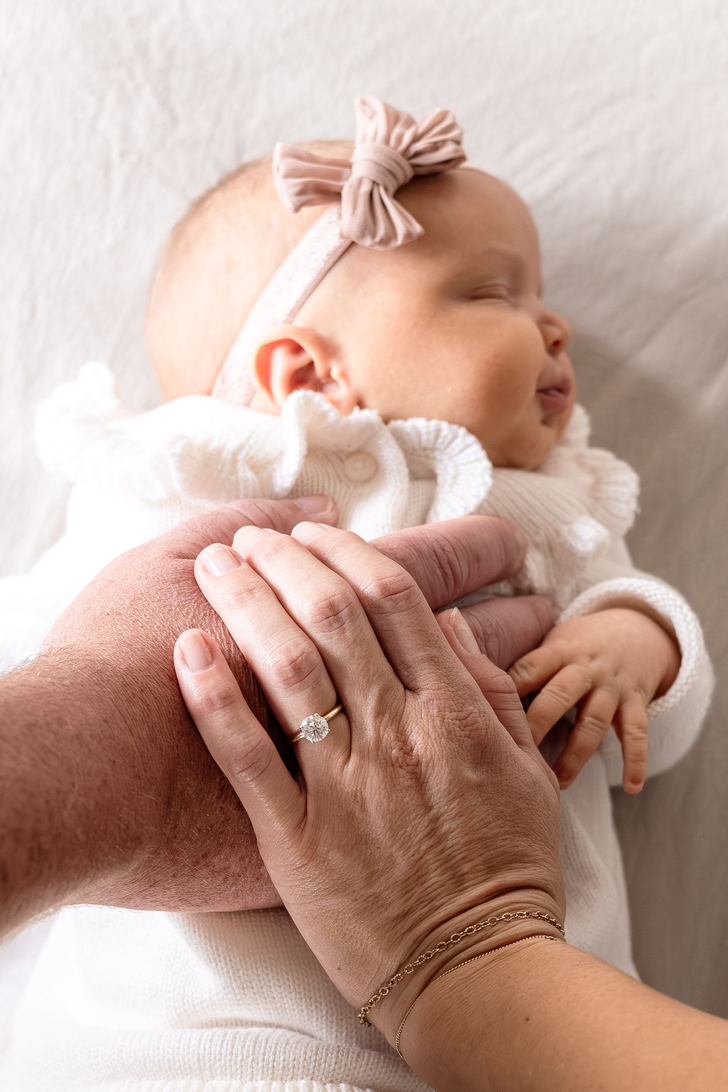 A series of gentle hands holding and comforting a newborn baby wearing white against soft white bedding.
