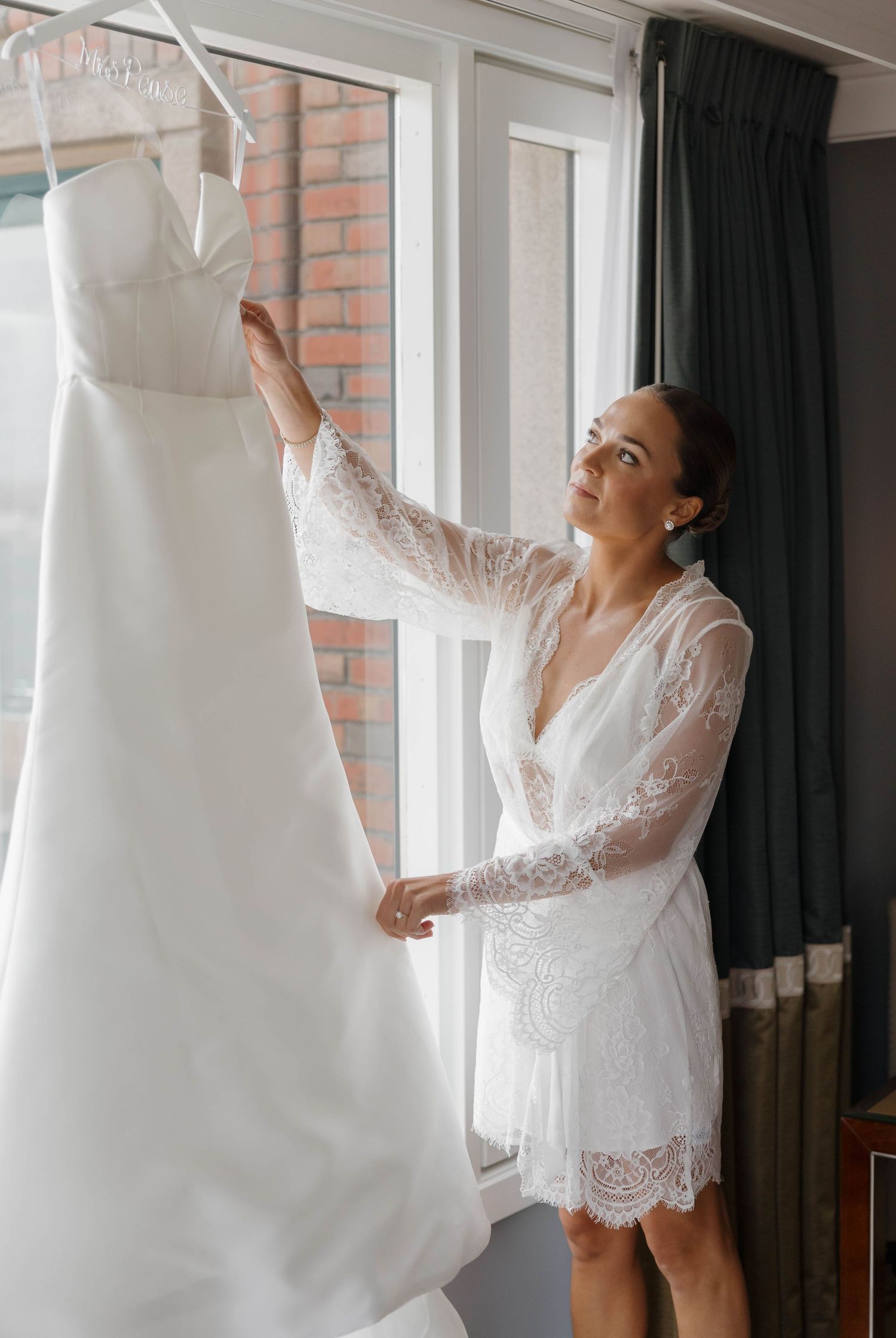 A white wedding dress hangs in a room while someone in a lace robe reaches up to touch it.