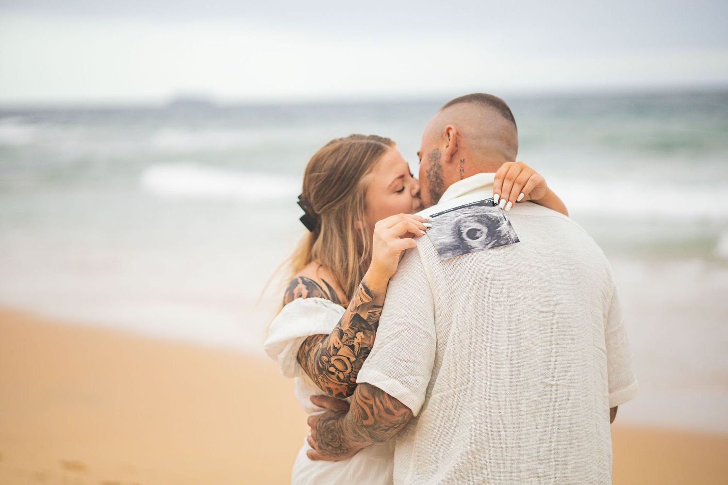 Couple embracing on beach holding ultrasound photo announcing pregnancy.
