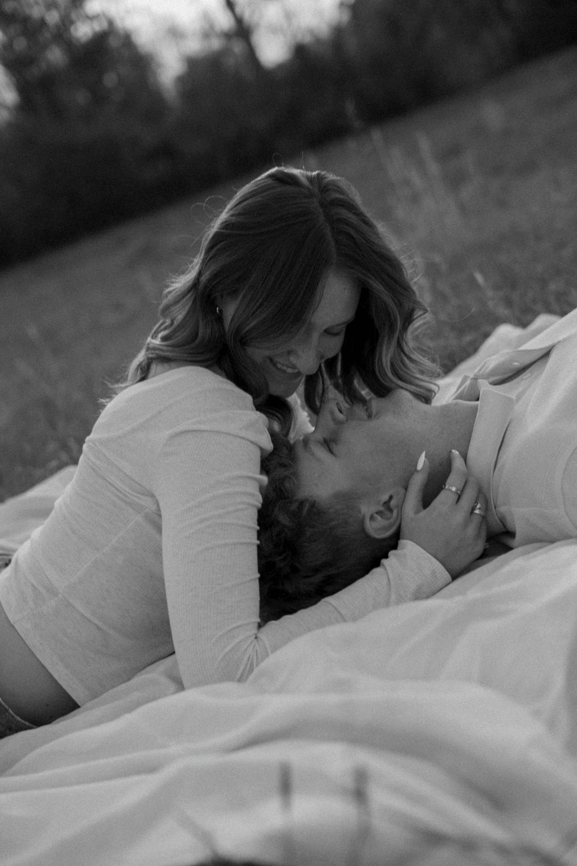 Black and white photo of a couple sharing a kiss while lying on a blanket in an open field.
