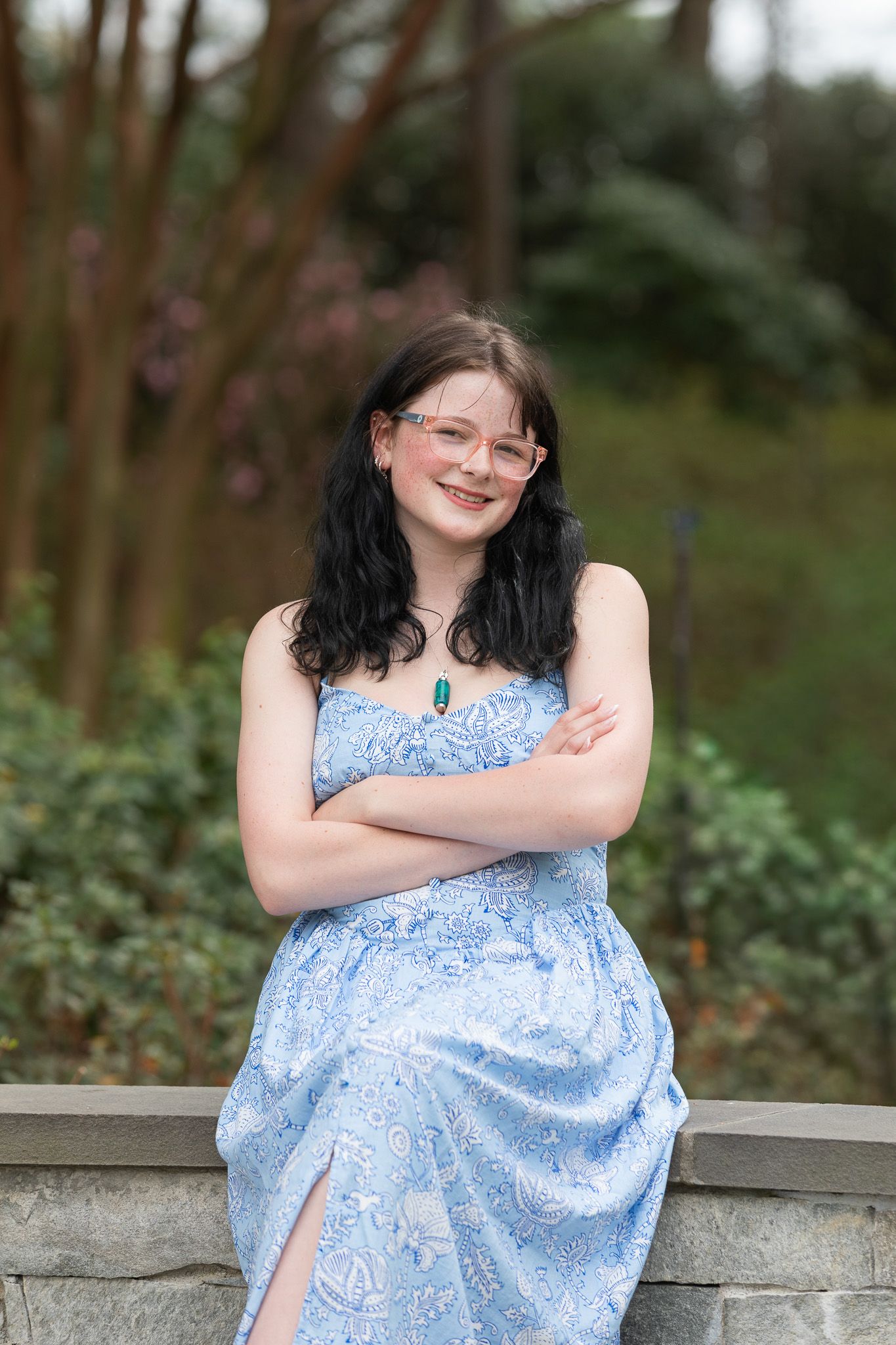 Young woman in blue paisley sundress with pink glasses and teal pendant necklace, smiling outdoors with arms crossed at WRAL Azalea Garden