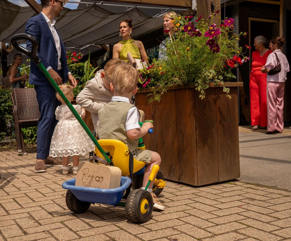Young boy riding a yellow and blue tricycle at an outdoor event with adults and flower decorations nearby.