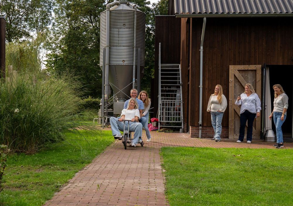 Een groep vrouwen loopt over een bakstenen pad in de buurt van een rustieke schuur met een graansilo op een groen boerenterrein. familie