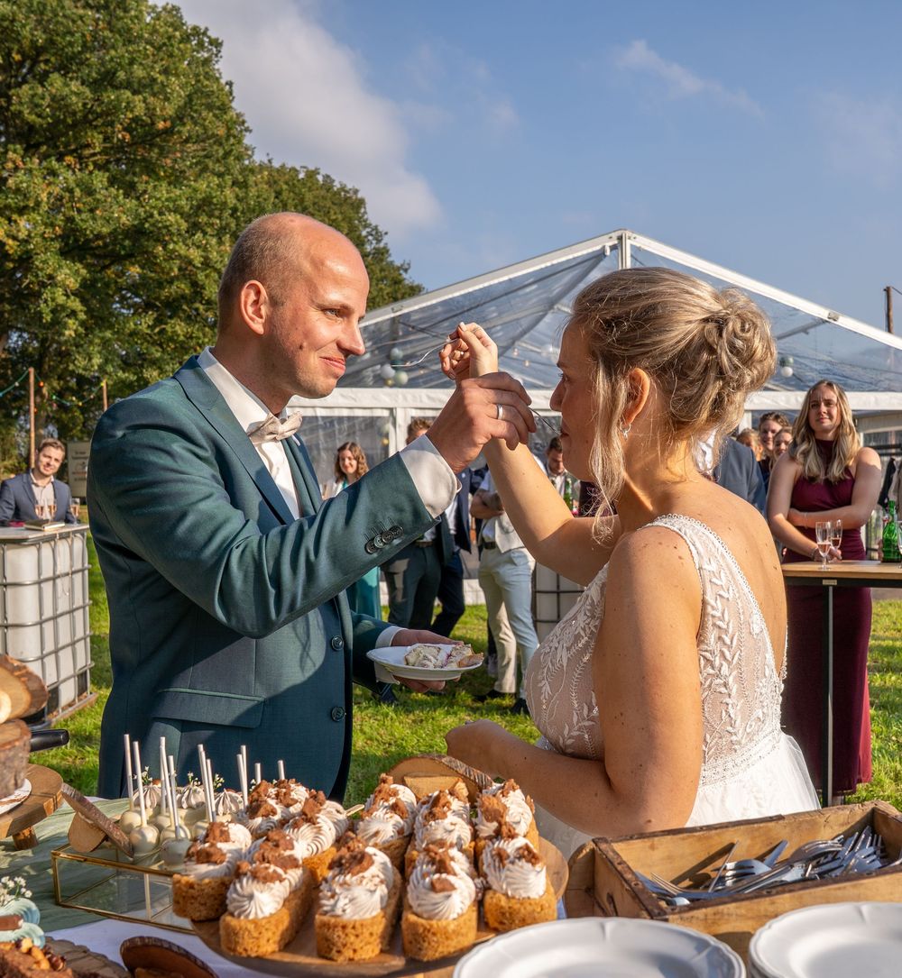 Bruid en bruidegom delen de bruidstaart tijdens een receptie in de buitenlucht, omringd door gasten en cupcakes.