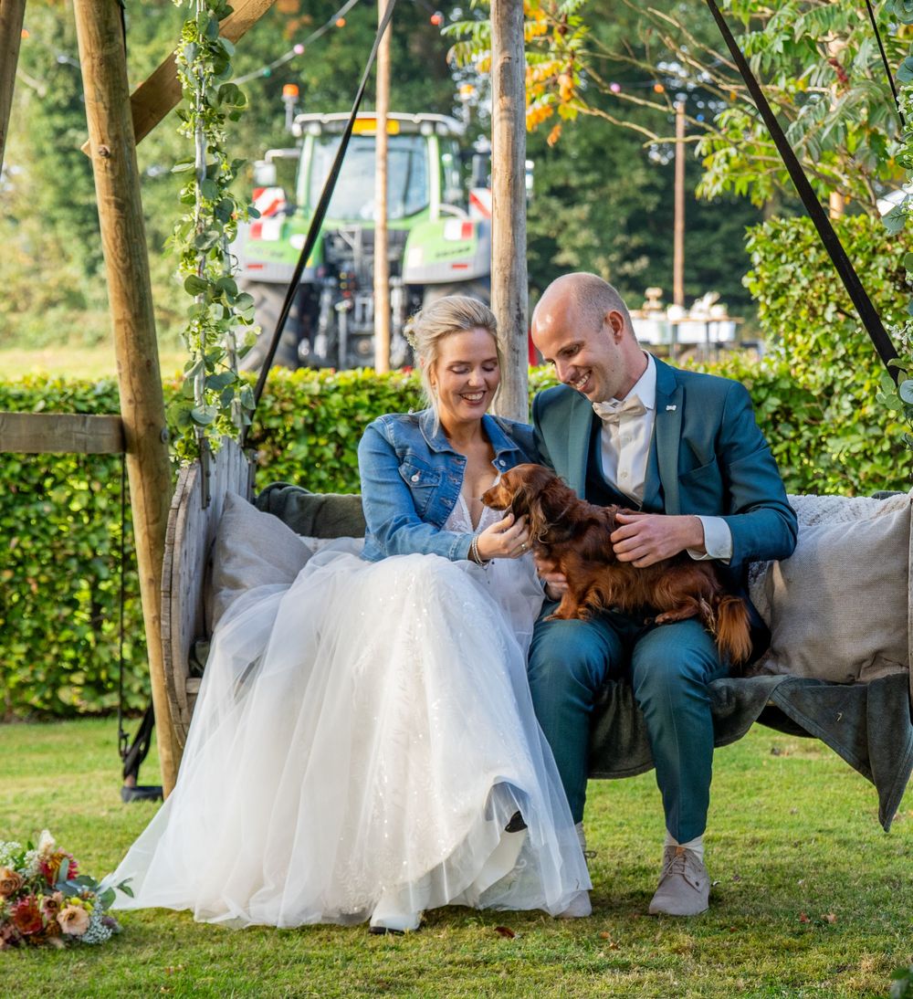 Bruid en bruidegom lachen samen op een schommel in de tuin, met een teckel in hun armen en een tractor op de achtergrond.
