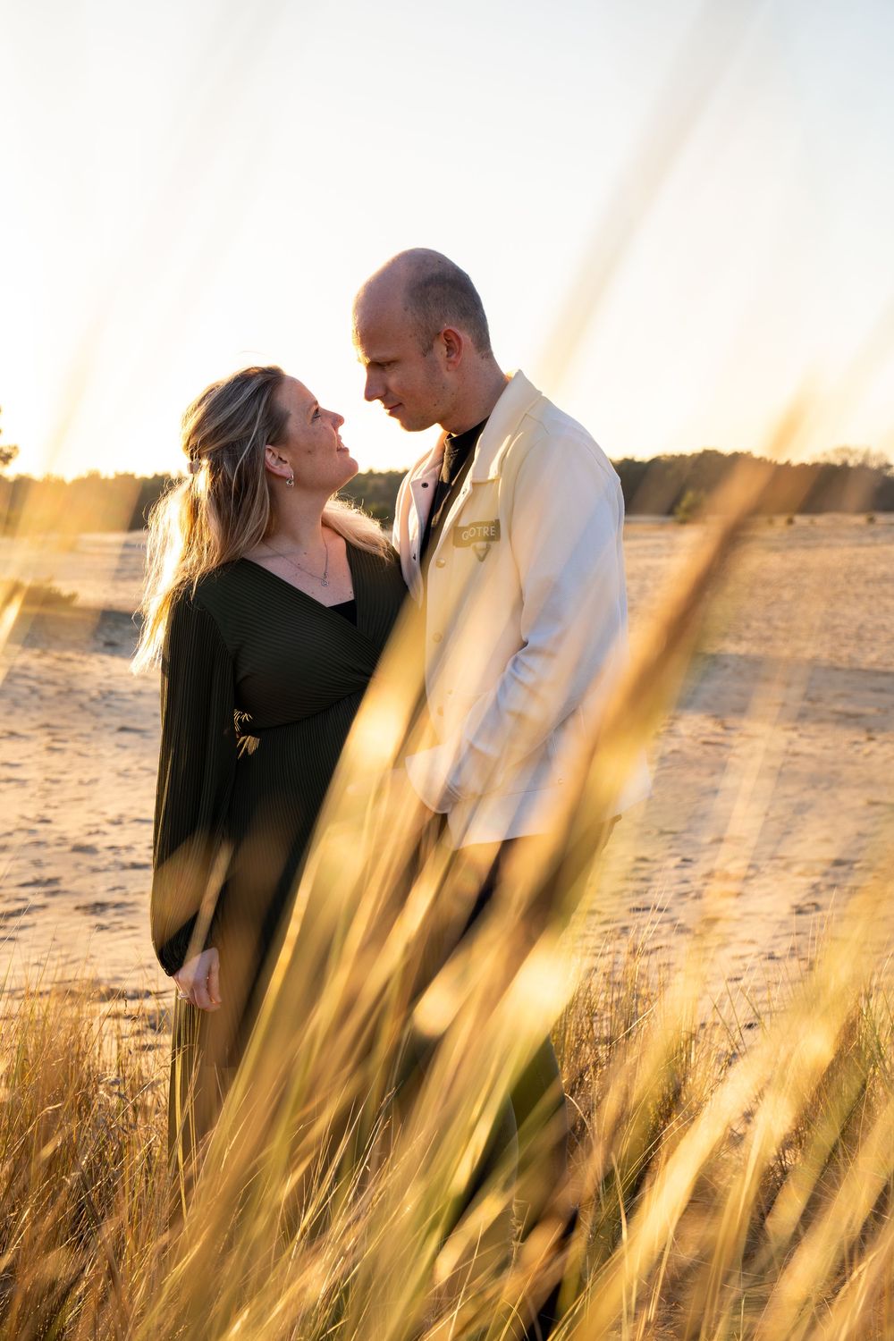 Een zwanger koppel deelt een intiem moment in het gouden strandgras tijdens een adembenemende zonsondergang.