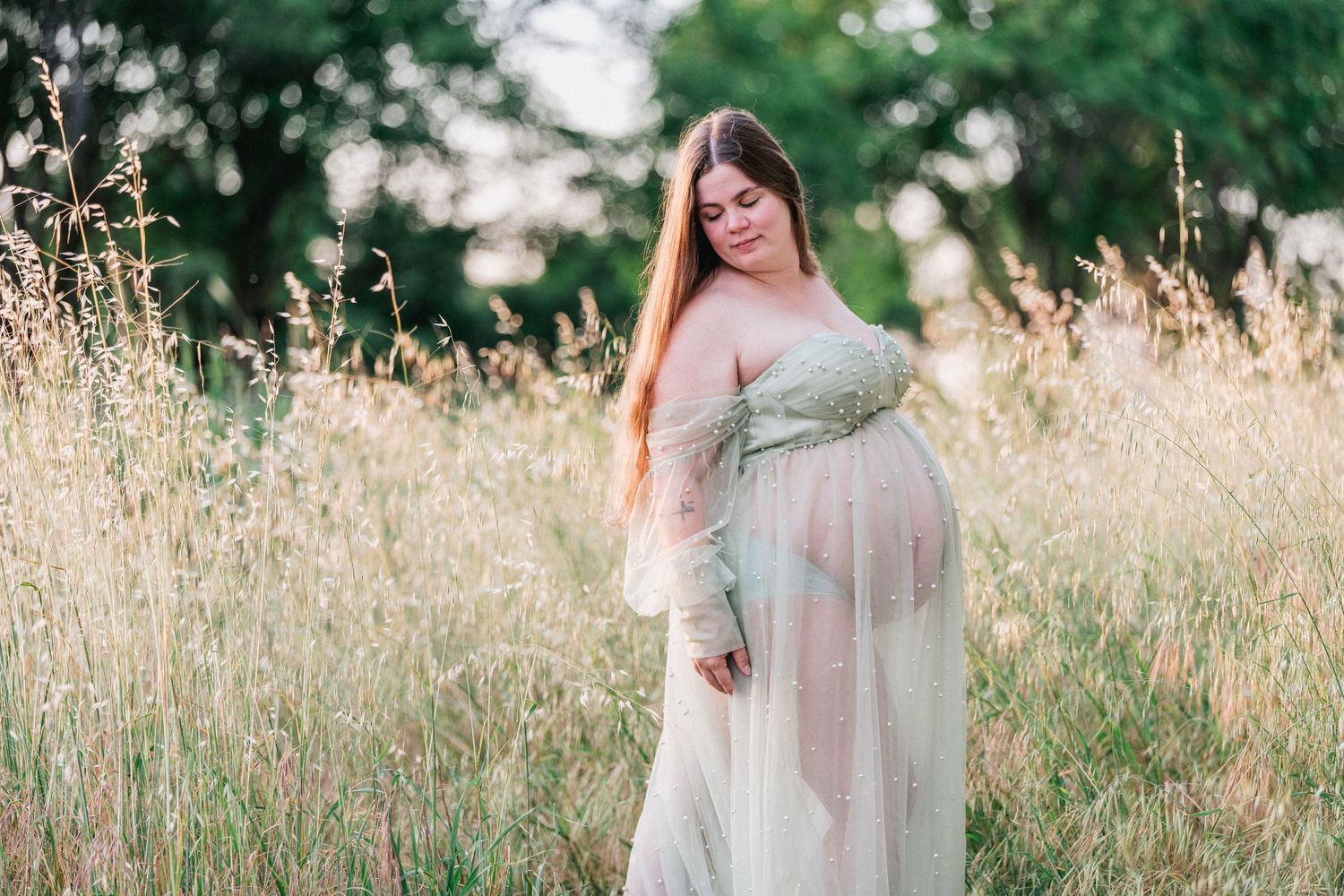 Pregnant woman in flowing green gown standing in golden meadow grass during golden hour maternity photo session.