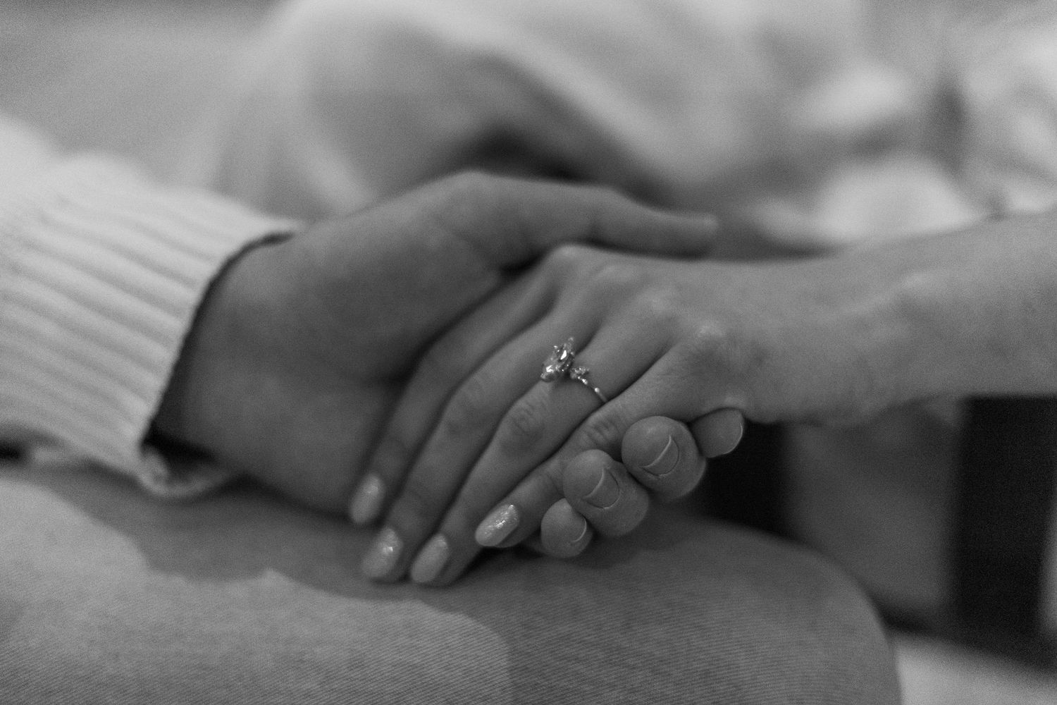 Black and white photo of two people holding hands, one wearing a diamond engagement ring, symbolizing love and commitment.