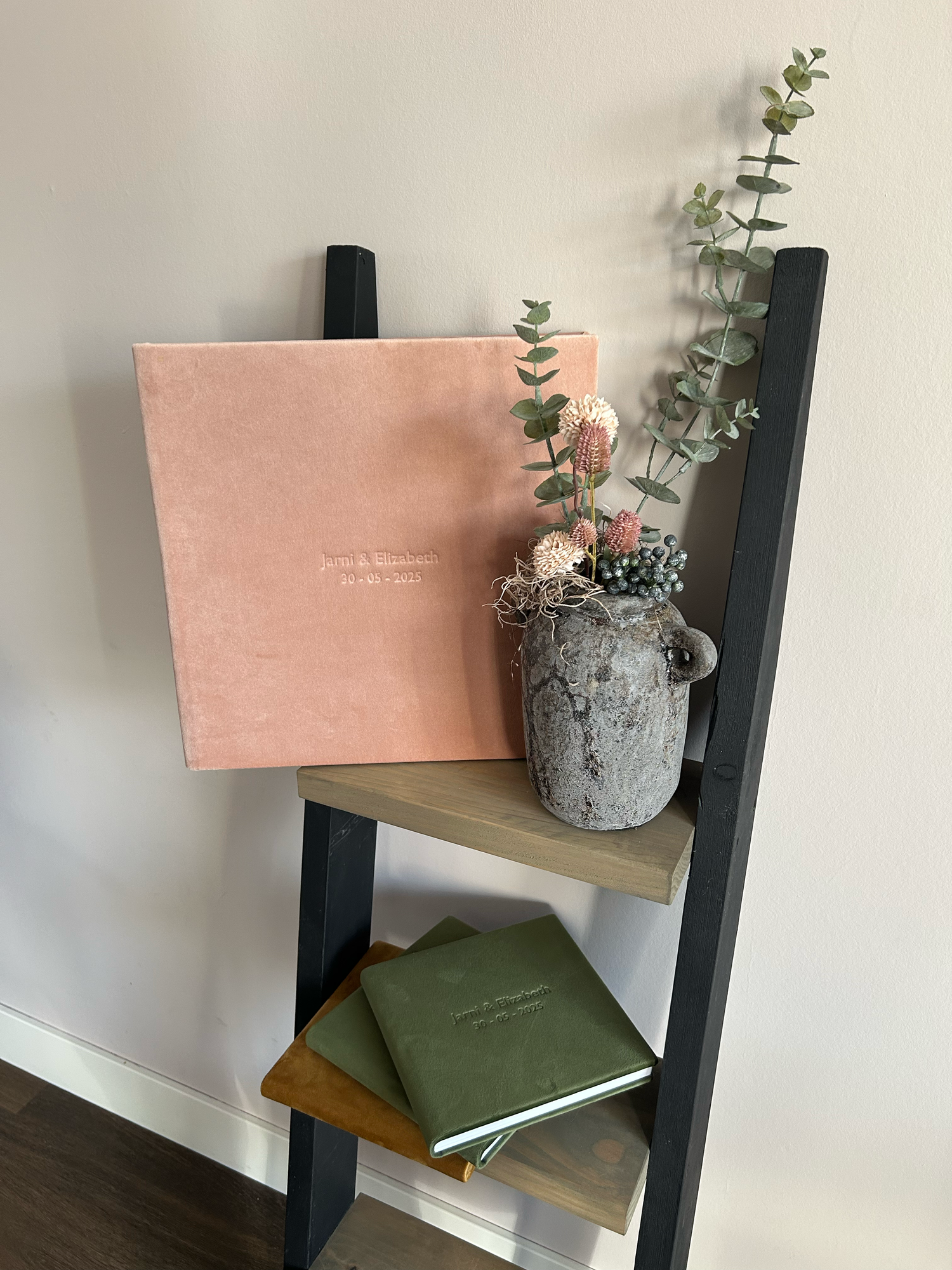 Roze fluwelen trouwalbum op een zwarte ladderplank met groene eucalyptus en gedroogde bloemen in een stenen vaas.