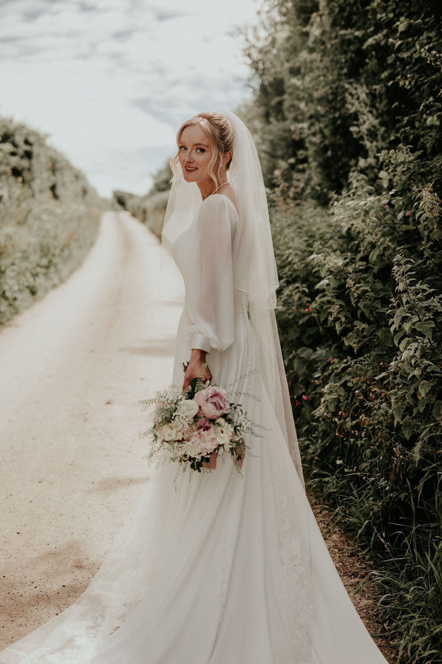 Smiling bride in off-shoulder lace wedding gown with veil holds bouquet, posing indoors with rustic wooden chairs.