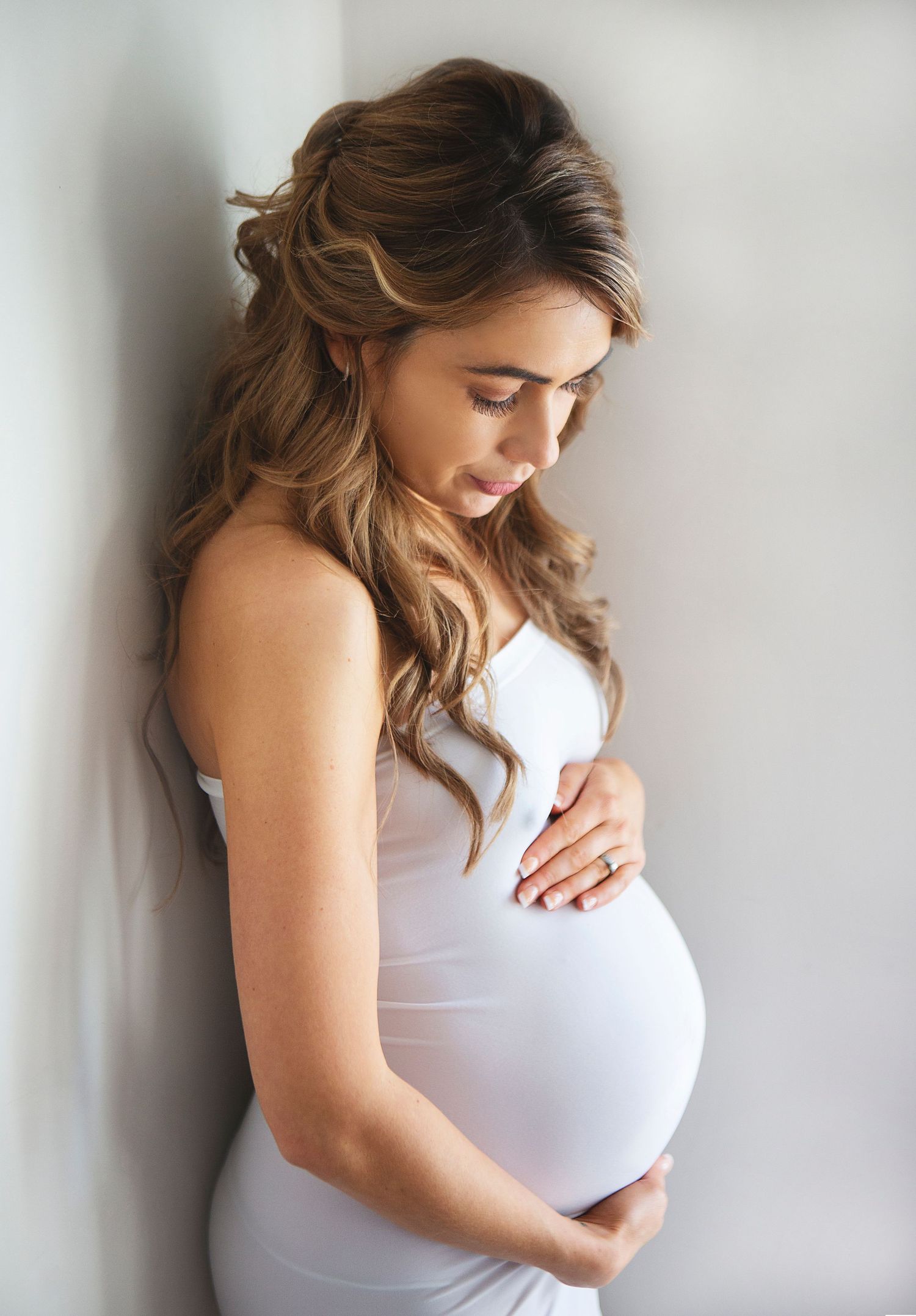 A pregnant woman in a white dress stands against a light wall in a series of artistic maternity photos. During a maternity session in Bexley