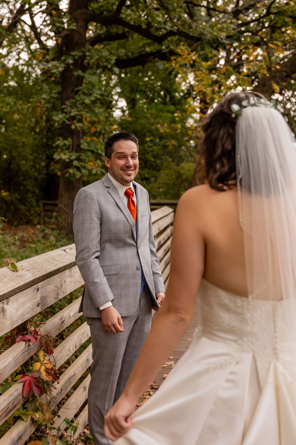 Groom in gray suit smiles at bride during first look moment on wooden bridge at autumn outdoor wedding at Platte River State Park
