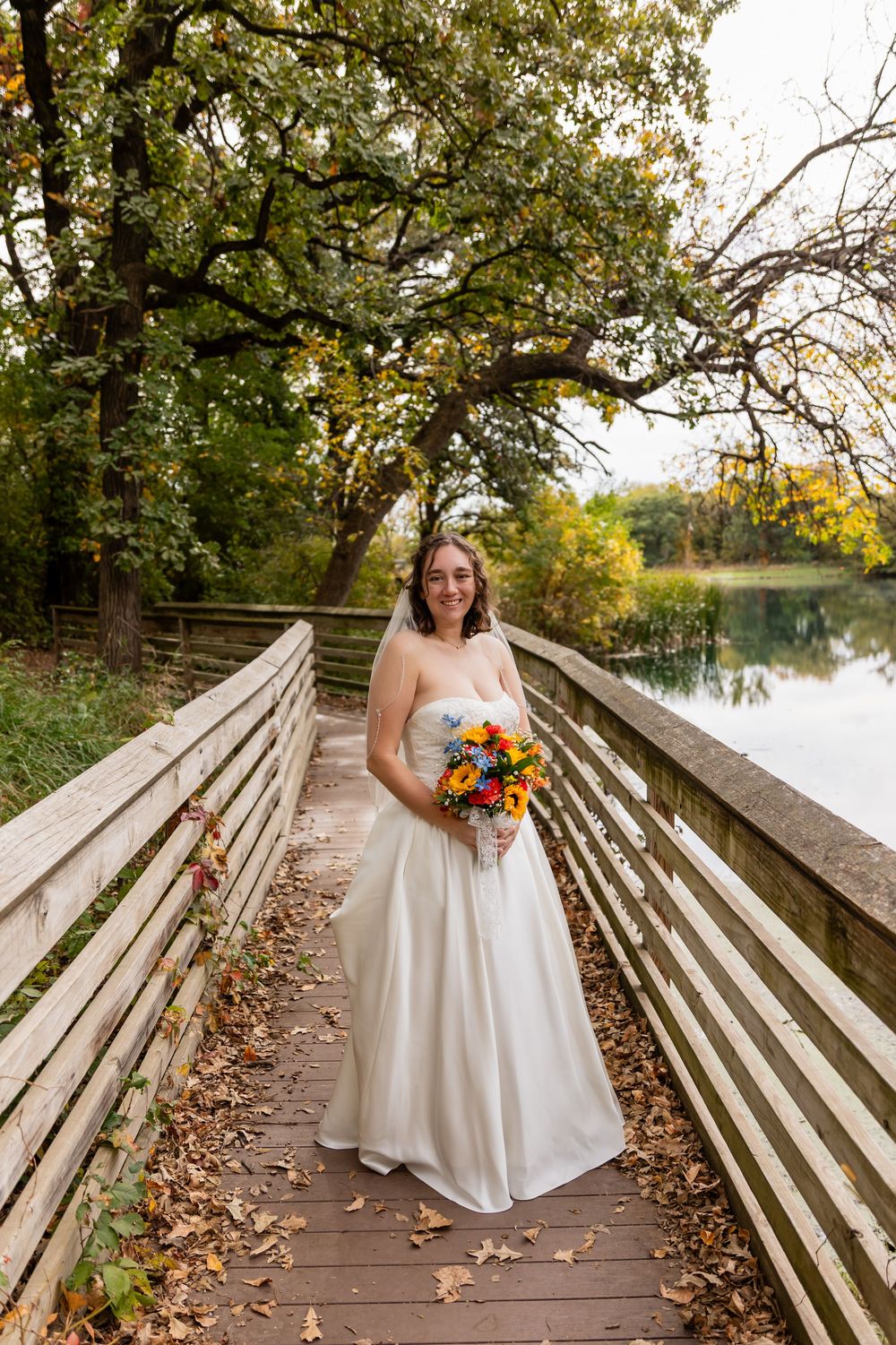 Bride in white strapless gown holding colorful bouquet stands on wooden bridge surrounded by autumn foliage at Platte River State Park
