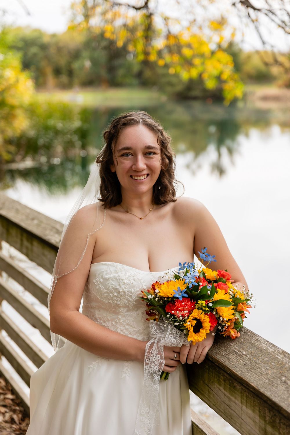 Smiling bride in strapless lace gown holding vibrant sunflower bouquet beside a serene lake in autumn at Platte River State Park