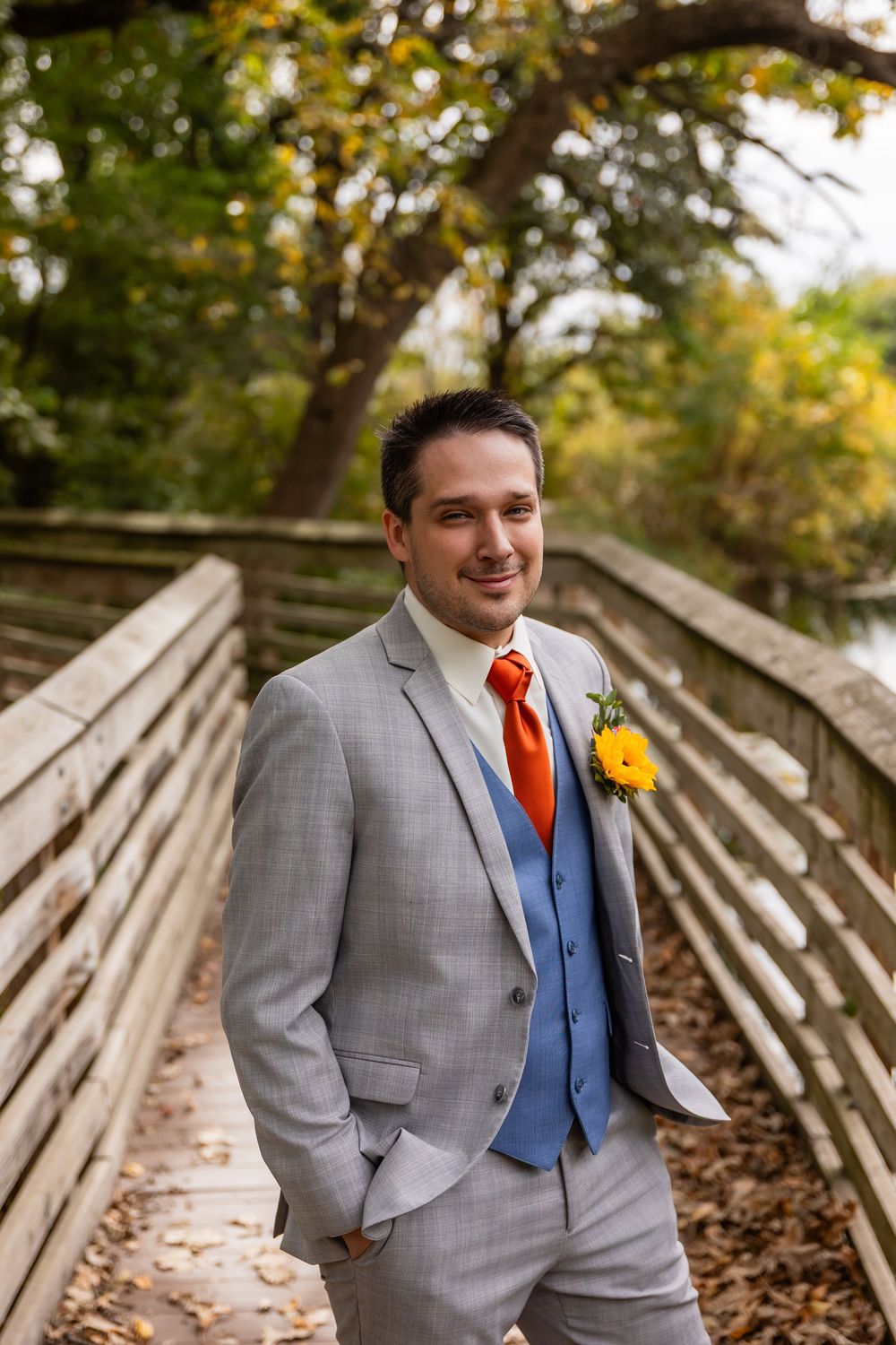 Groom in gray suit with orange tie and yellow boutonniere smiling on wooden bridge surrounded by autumn foliage at Platte River State Park