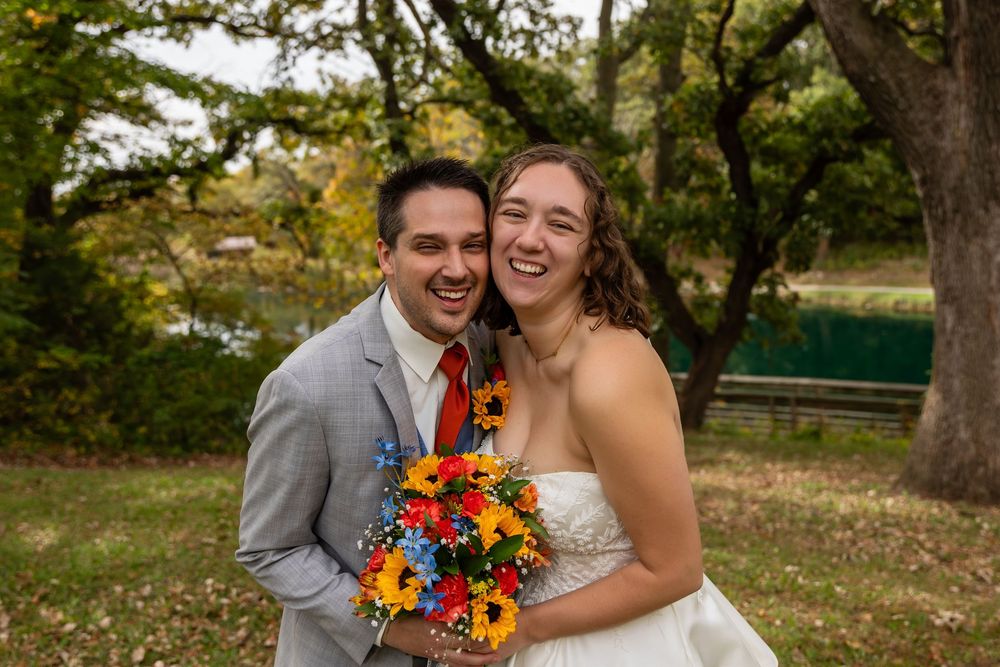 Happy bride and groom laugh together holding a vibrant sunflower bouquet outdoors at their fall wedding at Platte River State Park