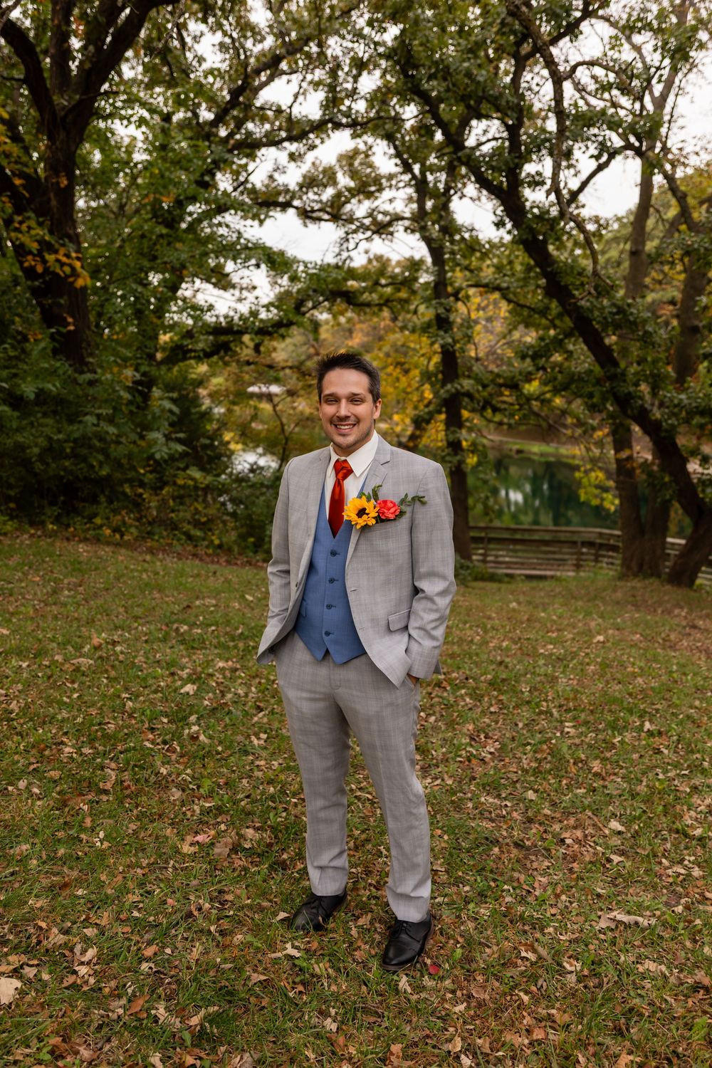 Smiling groom in light gray three-piece suit with red tie and colorful boutonniere posing outdoors in autumn park at Platte River State Park