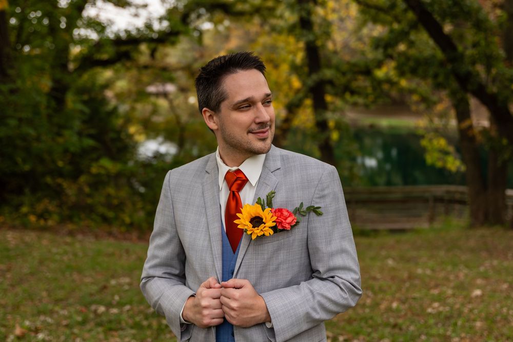 Groom in gray suit with red tie and sunflower boutonniere smiling outdoors among autumn trees at Platte River State Park in Louisville, NE