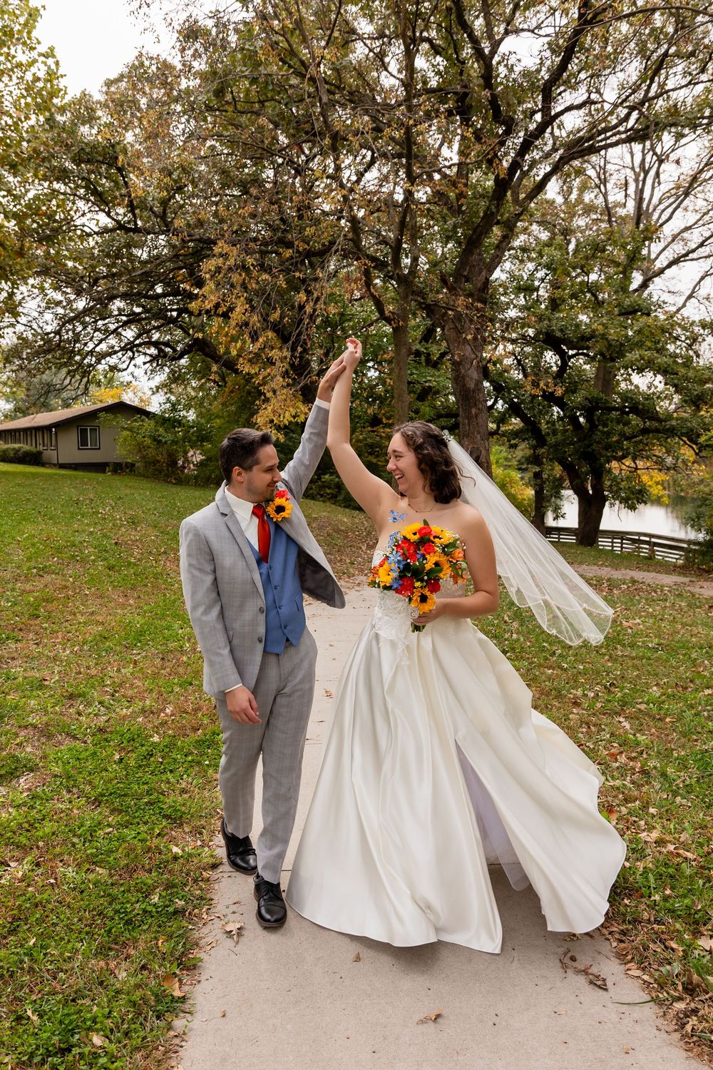 Joyful bride and groom dance on path, bride in white gown holding colorful bouquet amid fall trees at Platte River State Park in Louisville