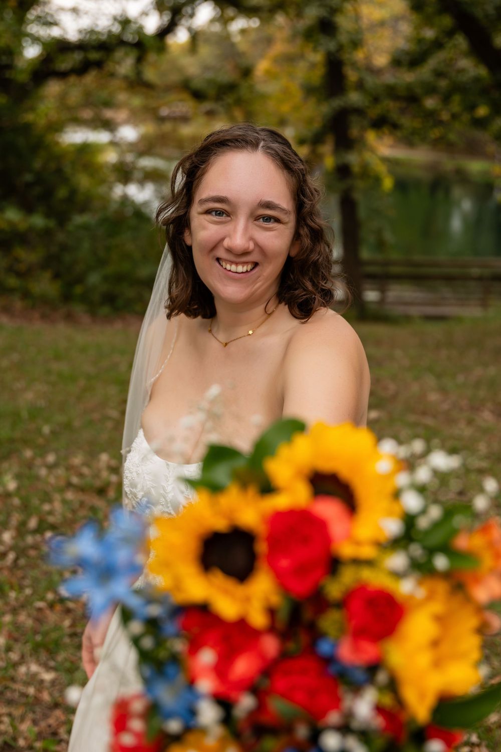 Smiling bride in white gown holding vibrant bouquet of sunflowers and colorful blooms in an outdoor autumn setting.