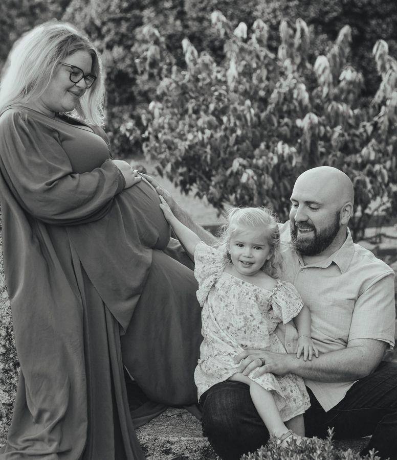 Black and white maternity photo of pregnant woman, bald man, and toddler girl outdoors among trees.