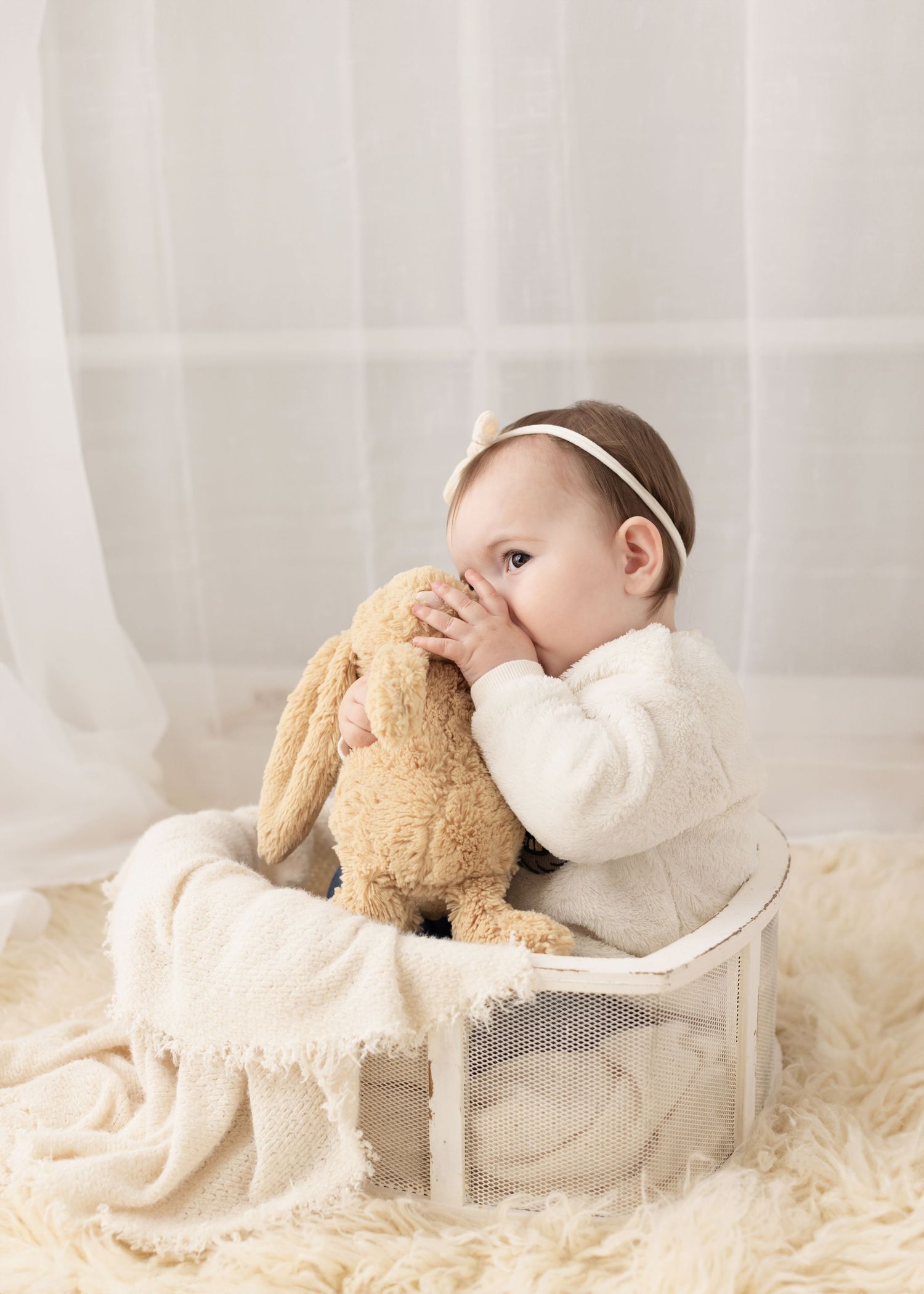 Baby in white clothing lies on cream textured bedding while playing with a soft brown teddy bear. Elizabeth Lang Photography Surrey