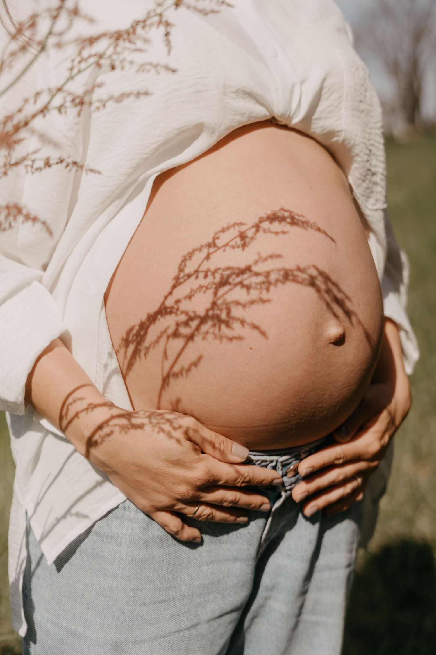 A pregnant woman in a white button-down shirt stands on a dirt path during an overcast day.