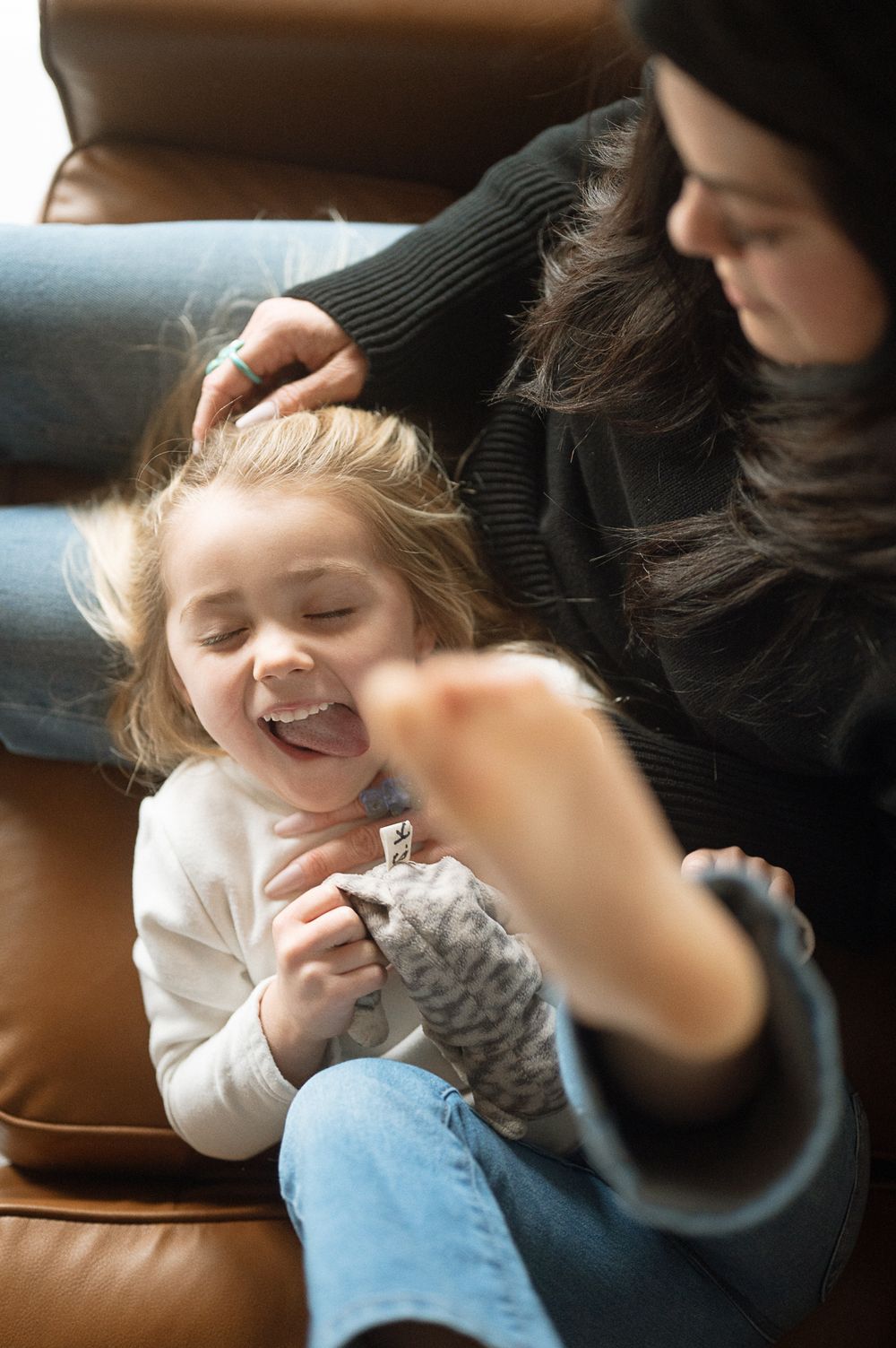 A playful moment shows a child and parent sharing laughter while relaxing on a brown leather couch.