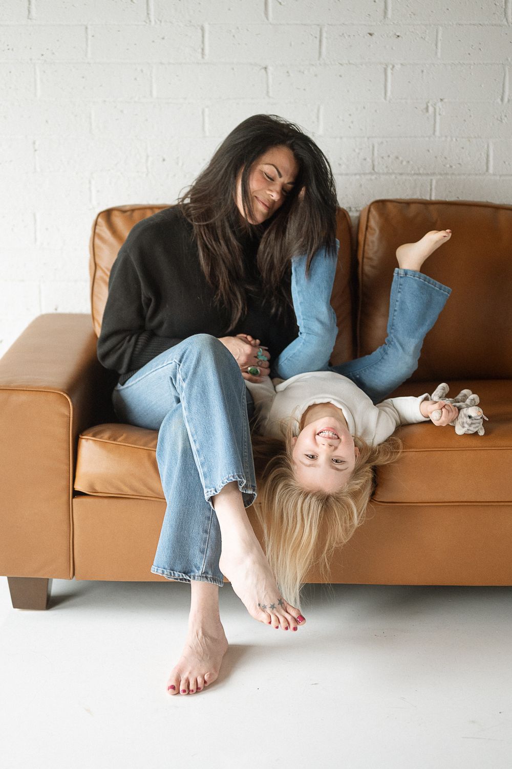 Two people in casual clothing relax playfully on a brown leather couch against a white wall.
