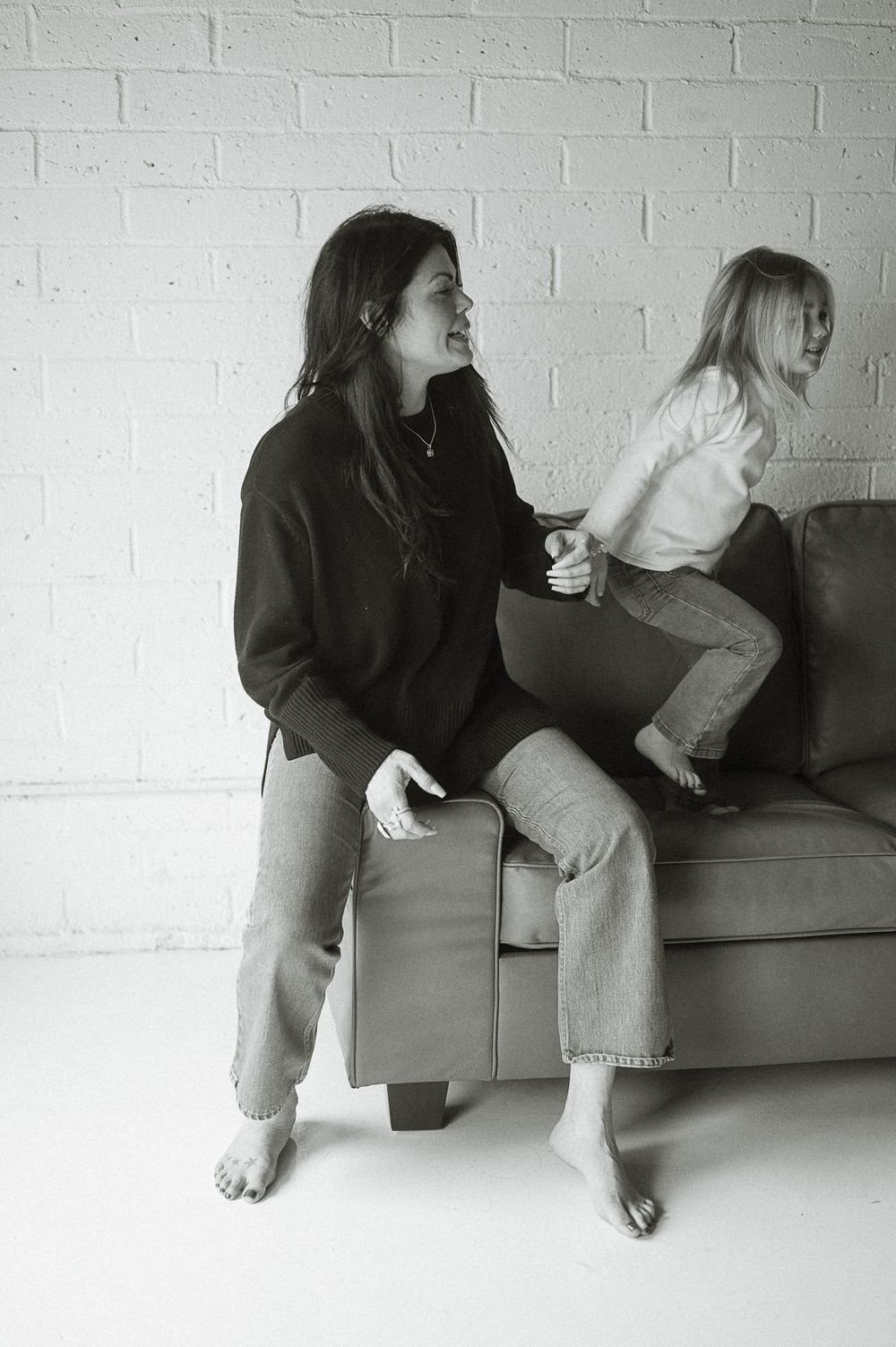 Two people in casual attire relaxing on a gray couch against a white brick wall in black and white photography.