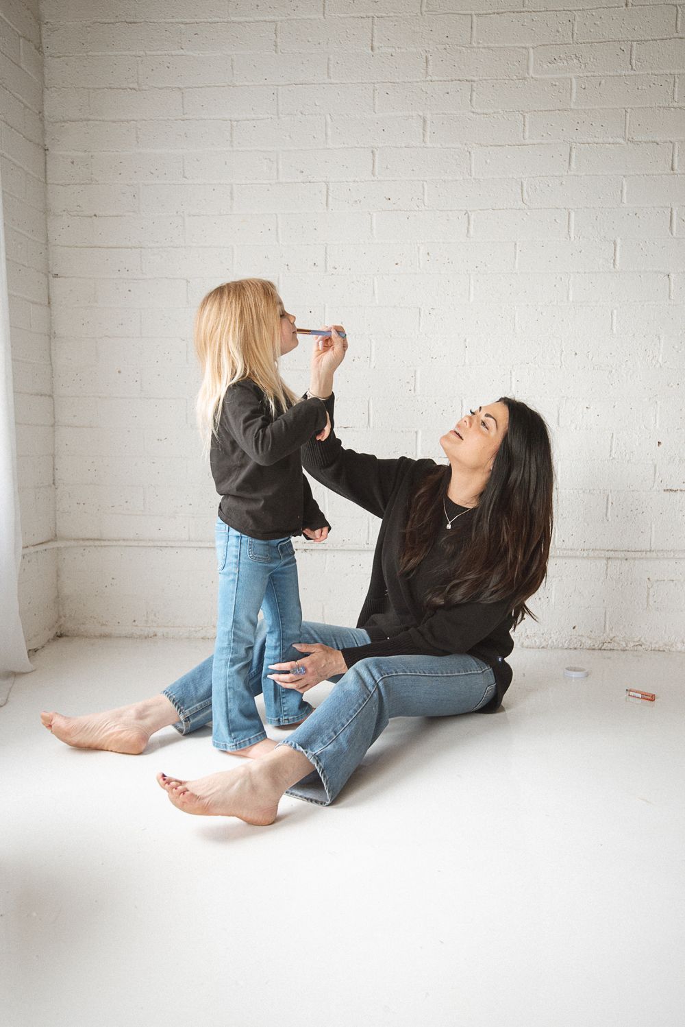 Two people in casual clothing playfully interacting on a white floor against a brick wall.