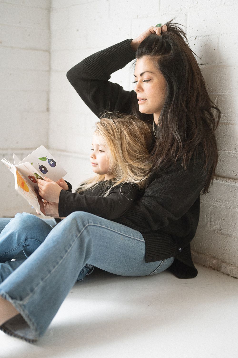 A person in jeans and black sweater lies curled up against a white wall reading a book in a minimalist setting.