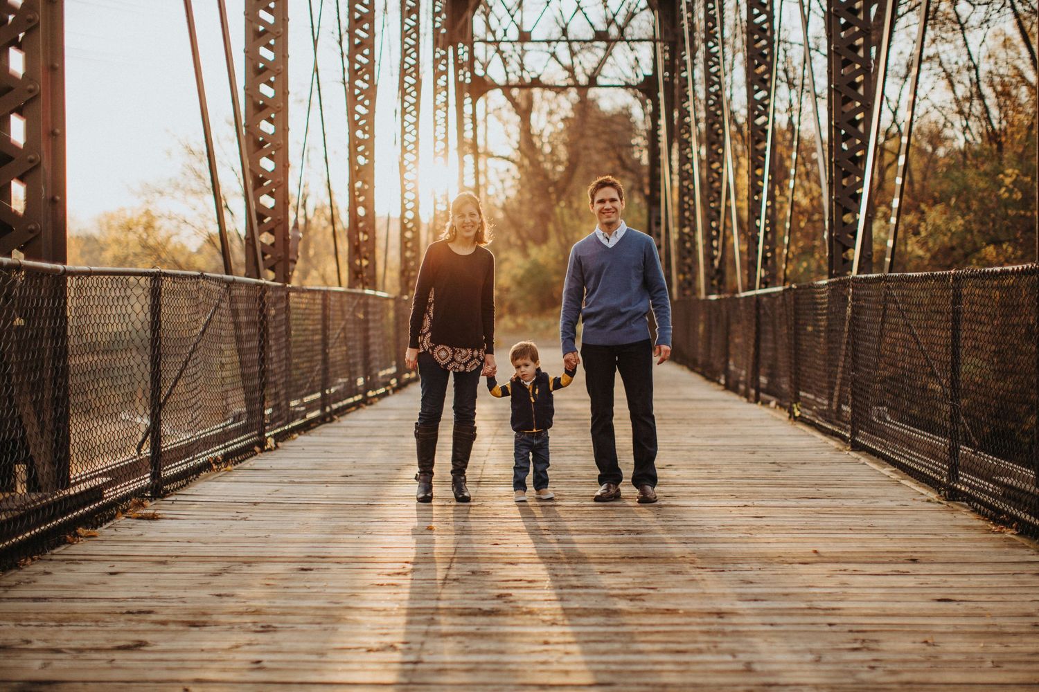 Three people walking together on a wooden bridge during golden hour sunset with metal railings and autumn trees.