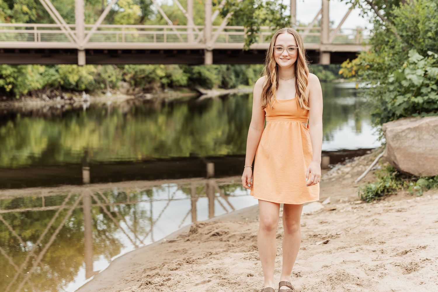 A person in a peach-colored dress stands on a riverbank near a wooden bridge on a sunny day.